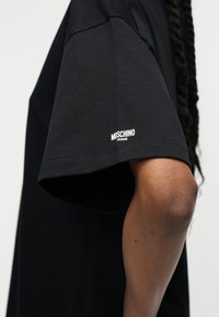 Άτομο που φοράει μαύρο T-shirt Moschino Jeans, με εμφανές βραχίονα και μανίκι που φέρουν το λευκό λογότυπο Moschino Jeans κοντά στο τελείωμα.