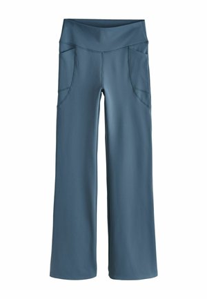 Vrouw met krullend blond haar die een zonnebril, sportbeha en broek draagt, steekt een straat over met een grote zwarte tas.