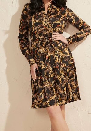 Kobieta ubrana w czarną sukienkę koszulową z żółto-brązowym wzorem łańcuchów, długimi rękawami, zapinaną na guziki z przodu, z paskiem przewiązanym w talii.