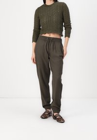 Vrouw in een olijfgroene gebreide cropped trui, bijpassende jogpants en luipaardprint ballerina's, staand tegen een simpele witte achtergrond.
