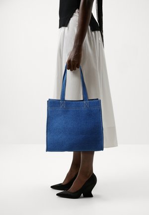 Personne portant une jupe blanche et un haut noir tenant un sac fourre-tout en jean bleu avec des poignées cousues, chaussée de chaussures noires à talons, sur fond blanc.