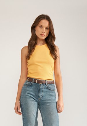 Femeie cu păr lung și castaniu, purtând un top galben fără mâneci, blugi albaștri cu talie înaltă și o curea maro, stând în picioare pe un fundal simplu.