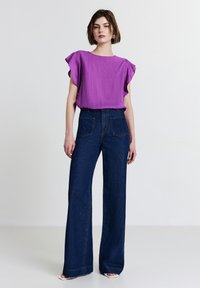 Donna in piedi che indossa una blusa viola a maniche corte, jeans a vita alta blu scuro a gamba larga e tacchi bianchi aperti sulla punta, su uno sfondo semplice.