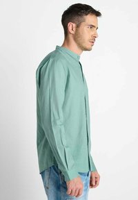 Man in een lichtgroen overhemd met mandarinekraag en lichtblauwe jeans, staand in profiel tegen een eenvoudige witte achtergrond.
