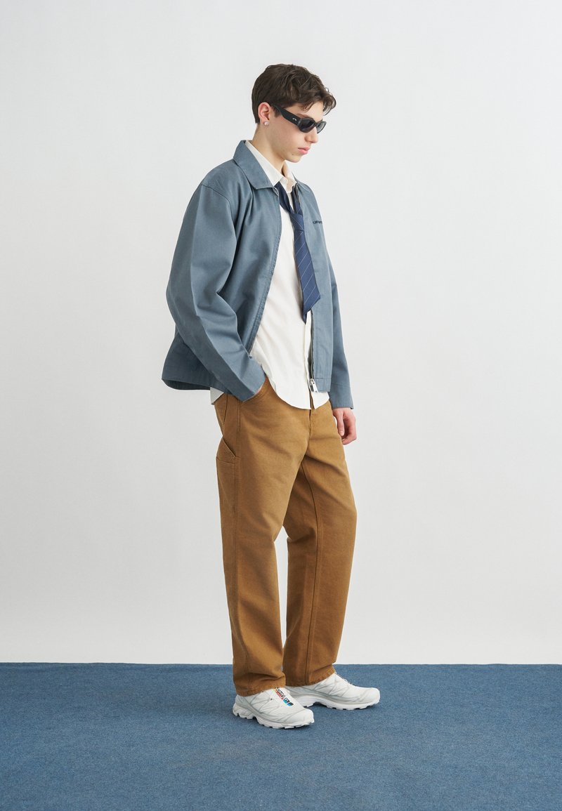 Jeune homme portant des lunettes de soleil, une veste bleue, une chemise blanche avec une cravate bleue lâche, un pantalon marron et des baskets blanches, debout sur un tapis bleu.