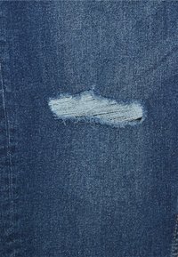 Tela de mezclilla en azul oscuro con una sección desgastada que presenta un desgarro horizontal deshilachado, exponiendo hilos de un azul más claro debajo.