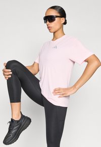 Ροζ αθλητική μπλούζα με κοντά μανίκια και γκρι λογότυπο, συνδυασμένη με μαύρο κολάν και αθλητικά μαύρα αθλητικά παπούτσια. Φοράει μεγάλα μαύρα γυαλιά ηλίου.