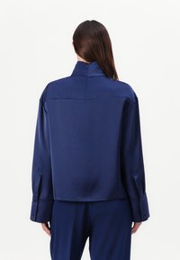 Camicia di raso blu navy con colletto alto, spalle scese e maniche oversize con dettagli a bottoni, che presenta una texture liscia.