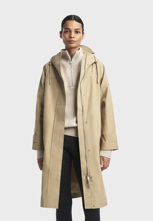 Kvinde iført en beige hættejakke over en lys beige quarter-zip sweater og mørke bukser, der står foran en ensfarvet baggrund.