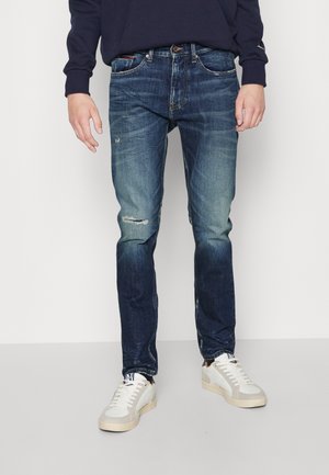 Mann trägt verblasste blaue Jeans mit leichter Abnutzung am einen Knie und weiße Freizeitsneaker, steht vor einem schlichten Hintergrund.