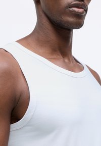 Balta bez piedurknēm tanks forma ar apaļu izgriezumu. Mīksta kokvilnas auduma virsma ir gluda un ar minimālu šuvju detaļu ap malām.