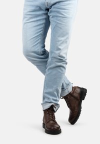 Persona que lleva puestos unos pantalones vaqueros azul claro y unas botas de cuero marrón oscuro con cordones, de pie con una pierna cruzada sobre la otra, sobre un fondo blanco.
