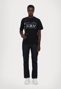 Black t-shirt with "ICE CREAM" text and skyline graphic, paired with dark jeans and white sneakers. Adjustable shoulder strap included.
