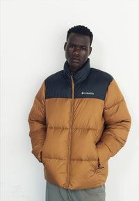 Doudoune marron et noire avec un col haut, fermeture zip, et logo sur la poitrine. Présente une texture matelassée et est isolée.