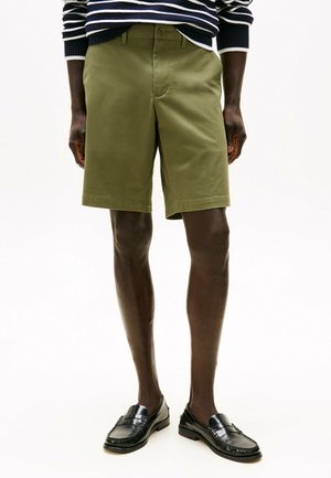Shorts verde oliva in cotone con una texture liscia, dotati di chiusura con bottone frontale e vestibilità casual. Abbinati a mocassini neri.
