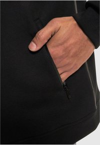 Sort sweatshirt med en lynlåslomme, glat tekstur, og et close-up af en hånd, der hviler i lommen, som fremhæver komfort og design.