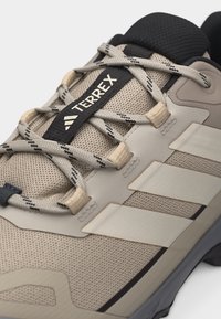 Κοντινό πλάνο μπεζ αθλητικού παπουτσιού Adidas Terrex με μαύρα και γκρι κορδόνια με σχέδιο, πλεκτό επάνω μέρος και τρεις χαρακτηριστικές ρίγες στο πλάι.