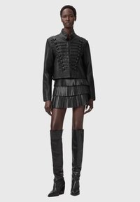 Μαύρο δερμάτινο κοντό μπουφάν με διακοσμητικά κουμπιά, συνδυασμένο με μαύρη δερμάτινη μίνι φούστα με πιέτες και μαύρες ψηλές μποτίνια με τακούνι.