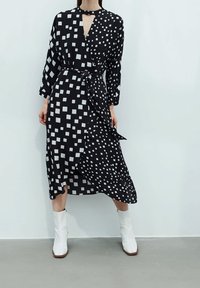 Schwarz-weißer gemusterter Wickelkleid mit langen Ärmeln, mit einem Schlüssellochausschnitt, einem Bindegürtel und einer fließenden Silhouette. Weiße Knöchelstiefel.