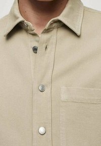 Camisa beige de botones con cuello clásico, botones plateados y un bolsillo en el pecho. La tela muestra una ligera textura.