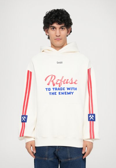 Witte hoodie met rode strepen, voorzien van blauwe en rode tekst met de boodschap "Weiger te handelen met de vijand" en een blauwe patch met een symbool op de mouw.
