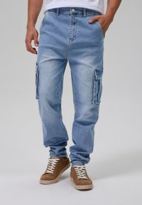 Helle blaue Denim-Cargohosen mit mehreren Seitentaschen, lockerer Passform und einer verwaschenen Textur. Tragen mit braunen Wildledersneakern.