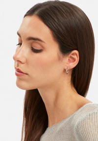 Orecchino a cerchio d'argento con design intrecciato, superficie riflettente e texture liscia, indossato sull'orecchio su uno sfondo neutro.