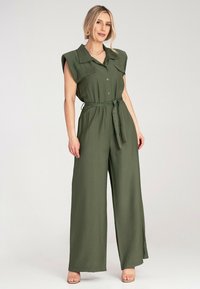 Kombinezon w oliwkowej zieleni z krótkimi, usztywnionymi rękawami, zapinany na guziki z przodu, z paskiem w talii. Wykonany z miękkiego, lekkiego materiału; szerokie nogawki.