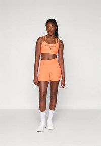 Fel oranje sportoutfit bestaande uit een sportbeha en shorts met hoge taille. Het materiaal lijkt rekbaar. Logo-accenten op beide stukken.