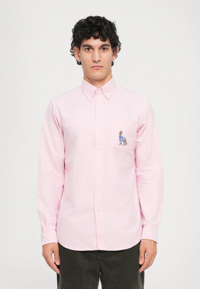 Camisa de botón rosa claro con mangas largas y un bolsillo en el pecho que presenta un diseño bordado. Tela de algodón suave con un cuello clásico.