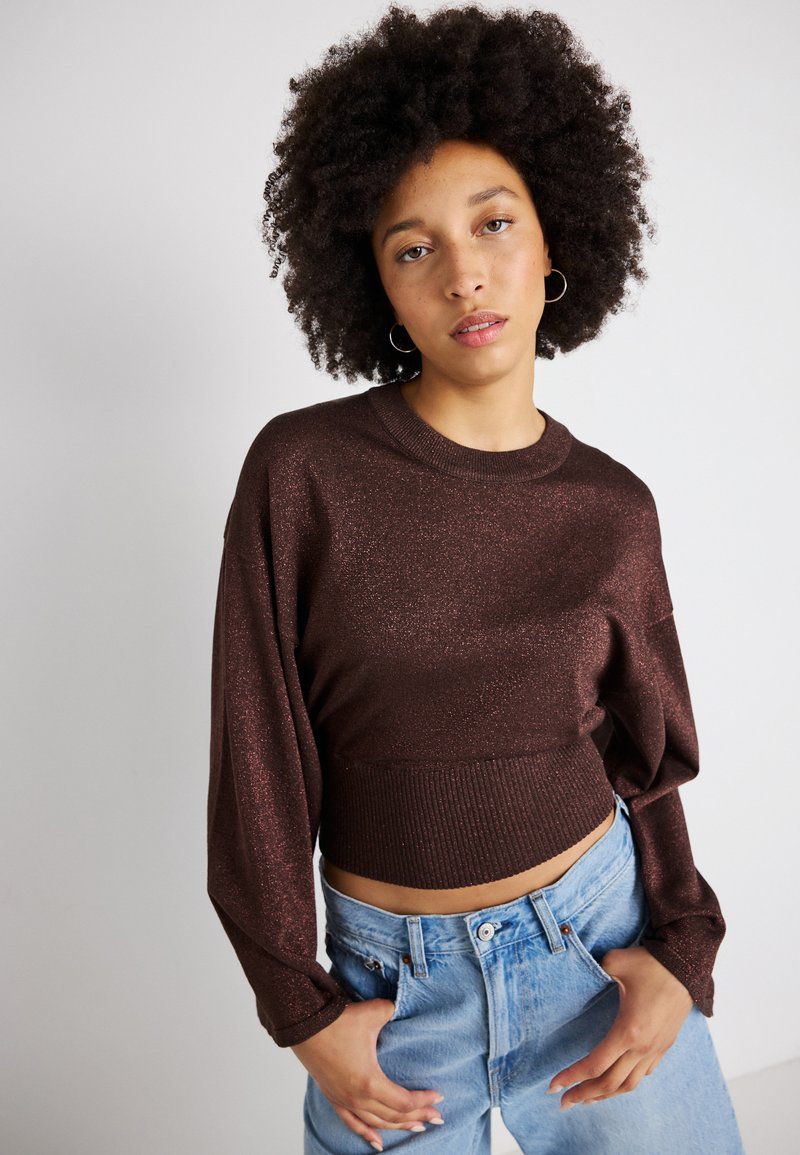 Suéter cropped castanho com brilho, de mangas longas e bainha canelada, combinado com jeans de ganga azul de cintura alta. Textura suave.