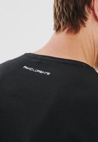 Czarna bawełniana koszulka z okrągłym dekoltem, z małym białym logo "PAKO LORENTE" na górze pleców. Miękka tekstura i casualowy styl.