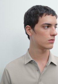 Polo shirt beige chiaro con colletto, realizzato in un tessuto testurizzato. Il modello ha i capelli corti e scuri e guarda di lato su uno sfondo neutro.