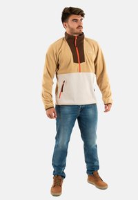 Chaqueta de forro polar en color marrón claro y blanco con detalles en marrón. Cuenta con un cuello alto, cierre de media cremallera en la parte frontal y dos bolsillos con cremallera. Se lleva con vaqueros.
