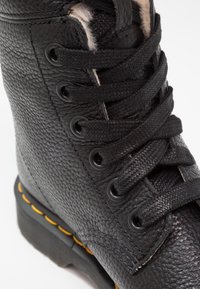 Botte en cuir noir avec une surface texturée, des lacets noirs ronds et des œillets en métal. Une couture jaune souligne la semelle pour créer un contraste.