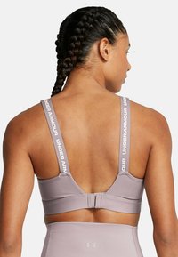 Sujetador deportivo Under Armour en lavanda clara; cuenta con tirantes ajustables con detalle del logotipo y un cierre de gancho en la parte posterior, acabado de tela suave.