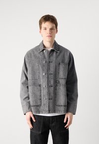 Grå denimjakke med kragehals, fire frontlommer og kontraststing. Båret over en hvit skjorte, med en avslappet passform.