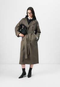 Oliivroheline trench-kate vöökohaga, topelt-rinnapartii nuppudega ja augukestega detailidega. Must mini käekott ja terava ninaga pahkluu saapad.