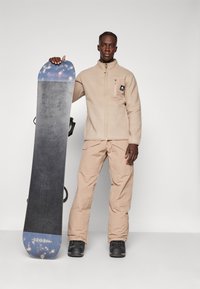 Beige fleece-jakke med høy krage, glidelåslomme; beige snøscooterbukser; snowboard med blå og svart design, med teksturert overflate.