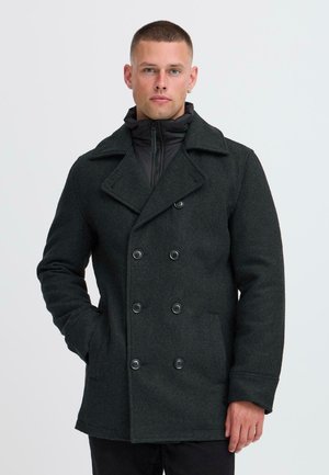 Dunkelgrauer Woll-Peacoat mit doppelter Knopfleiste, großen Knöpfen und seitlichen Taschen, über einer schwarzen Reißverschluss-Puffjacke getragen.