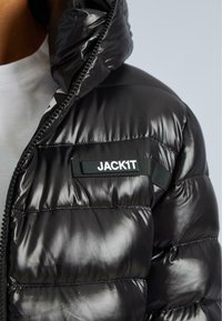 Mirdzošs melns quiltēts jaka ar augstu apkakli, ieģērbts ar daļēji atvērtu rāvējslēdzi, ar "JACK1T" emblēmu uz krūtīm, virs baltas kreklas.