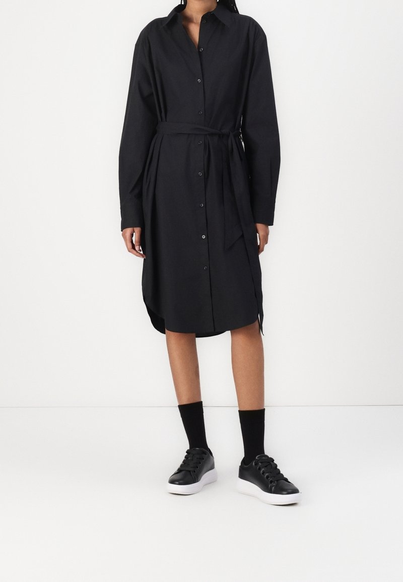 Femme portant une robe chemise noire à manches longues avec des boutons et une ceinture à la taille, des chaussettes noires et des baskets noires avec des semelles blanches.