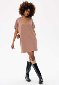 Swetrowa sukienka w delikatnym różowym kolorze z dekoltem w kształcie litery V, krótkimi rękawami i luźnym fasonem, zestawiona z wysokimi czarnymi butami i prążkowanymi kremowymi skarpetkami.