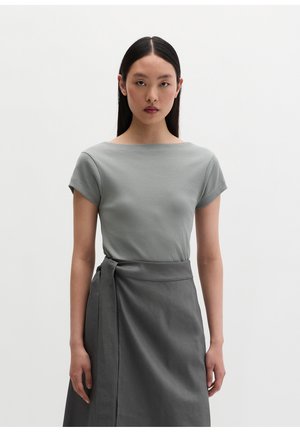 Femme aux longs cheveux noirs portant un haut gris à manches courtes et une jupe portefeuille gris foncé, debout devant un fond blanc uni.