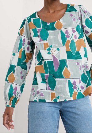 Woman wearing long-sleeve blouse with abstract colorful vase and flower pattern, paired with light blue jeans.