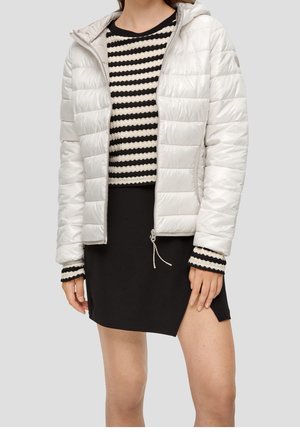 Vrouw draagt een witte gewatteerde jas met ritssluiting over een zwart-wit gestreepte trui en een zwarte rok met een voorin een split.
