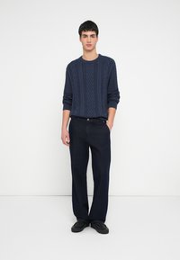 Granatowy sweter w warkoczowy splot z prążkowanymi mankietami i okrągłym dekoltem, w połączeniu z ciemnymi prostymi spodniami i czarnymi butami. Prosty, teksturowany wzór.