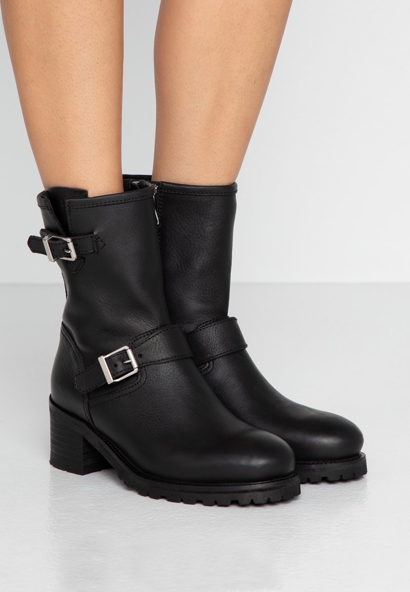 Černé kožené kotníkové boty s texturovaným povrchem, stříbrnými přezkami a nízkým blokovým podpatkem. Mají zaoblenou špičku a robustní podrážku.