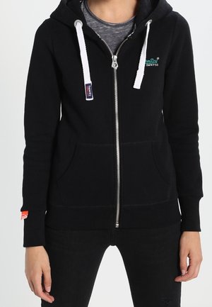 Osoba nosząca czarną bluzę z kapturem zapinaną na zamek, z białymi troczkami, przednimi kieszeniami i małym zielonym logo na lewej piersi.