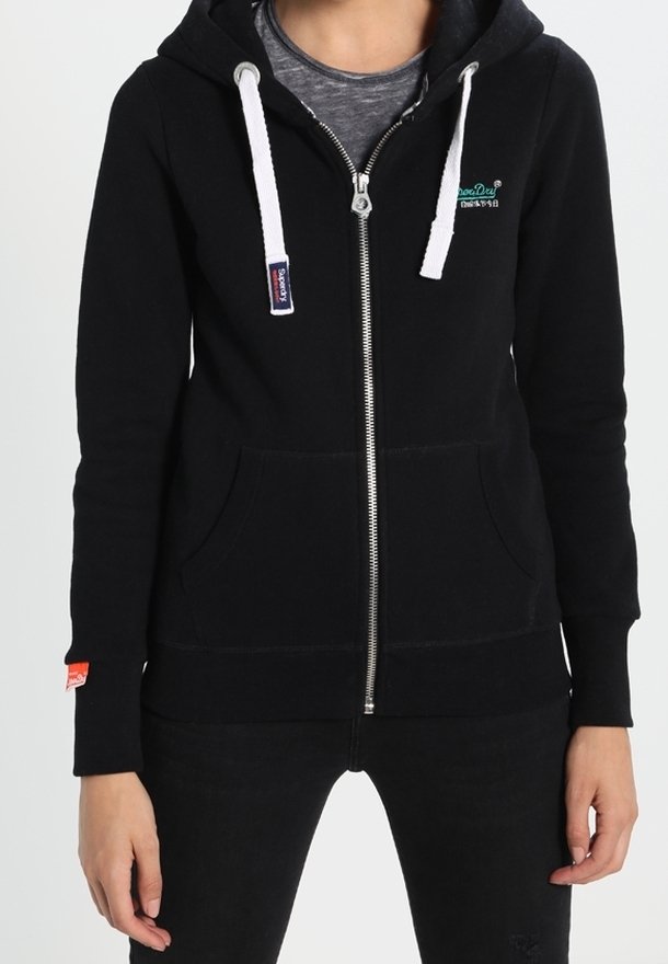 Personne portant un sweat à capuche noir zippé avec des cordons blancs, des poches avant et un petit logo vert sur le côté gauche de la poitrine.